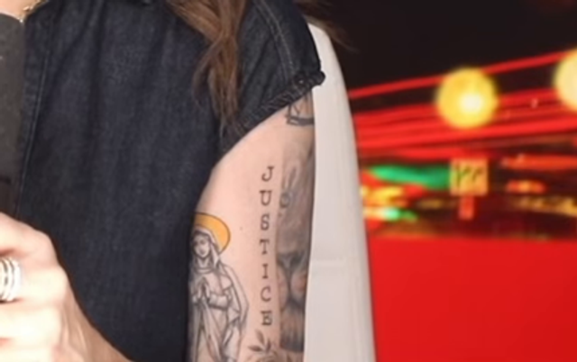 Close-up detail of the studio-desk forearm: a robed, haloed saint or Madonna figure beside vertical lettering that reads JUSTICE, with additional script and a large-cat face.