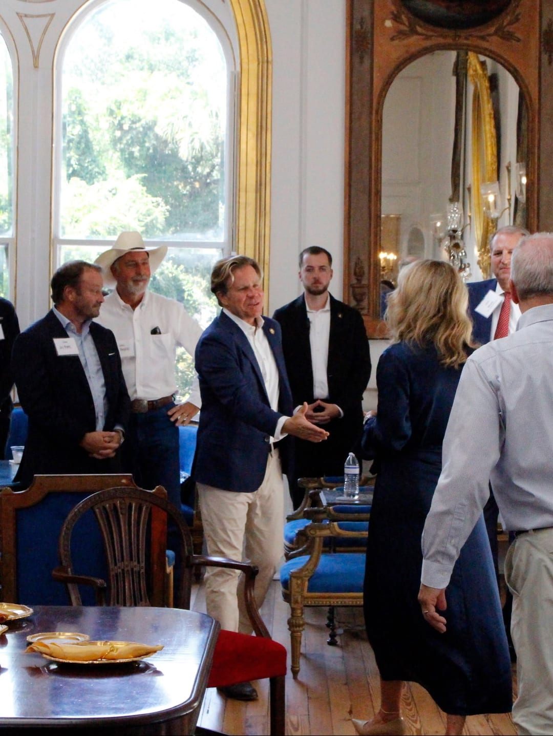 A crowded, gilded Charleston reception room; guests mingle around a table set with place cards.