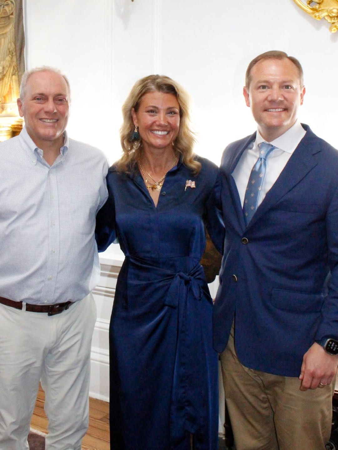 Republican congressional nominee Jenny Costa Honeycutt (center), in a navy dress with an American-flag lapel pin, stands between two male guests at an ornate Charleston reception. Her campaign identified the visiting guests as House Majority Leader Steve Scalise and Rep. Mike Haridopolos. Nancy Mace does not appear.