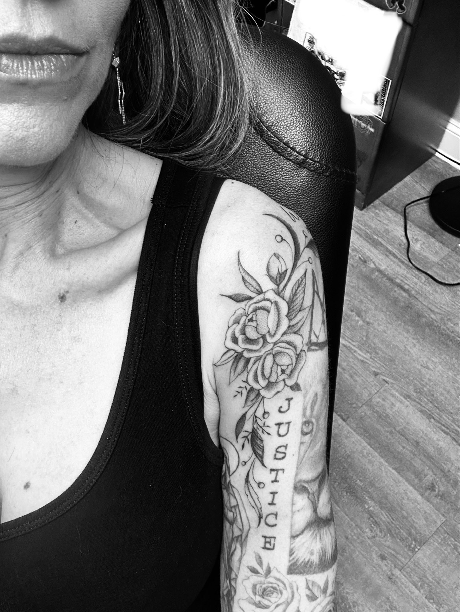 Black-and-grey photo of Nancy Mace's left upper arm in a tattoo chair: peonies and leaves with a crescent moon and geometric line work on the shoulder, the vertical typewriter lettering JUSTICE down the arm, a lion's face beside it, and roses below.