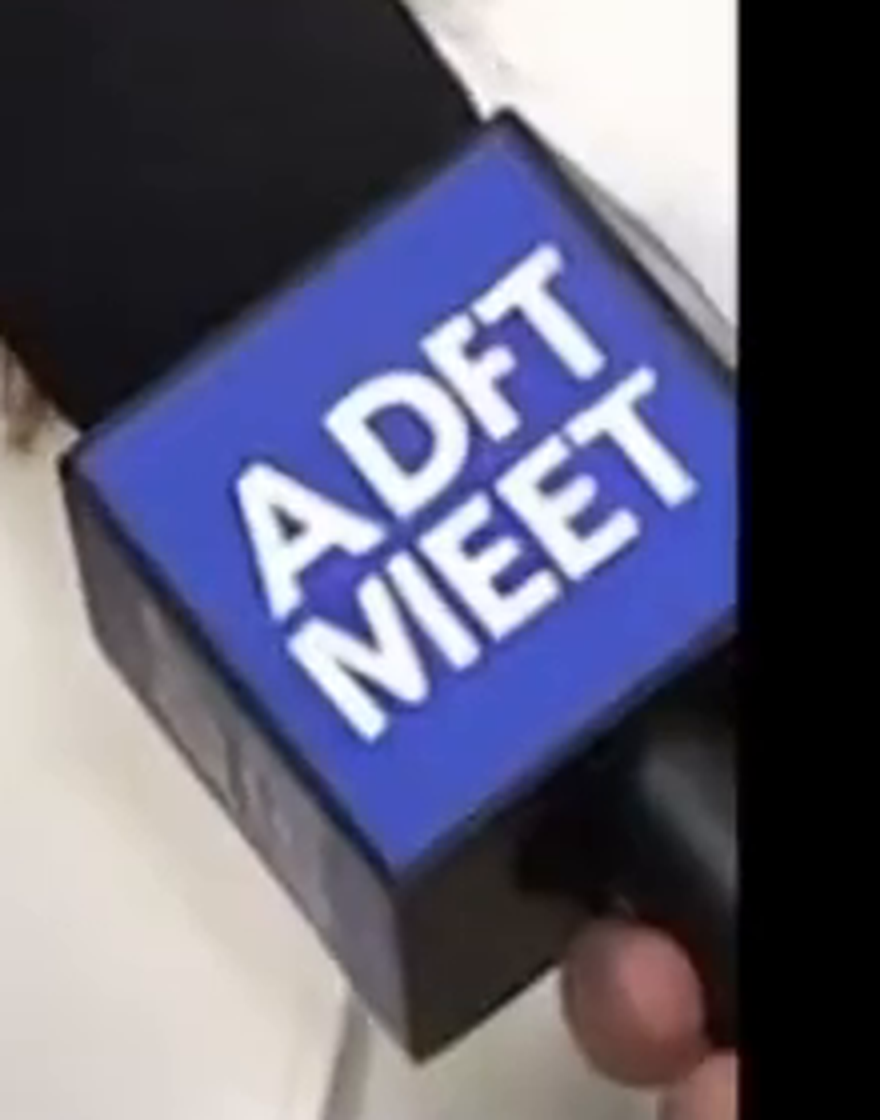 Zoomed crop of the right microphone flag from the GET MACED press-gaggle frame: a blue flag with white lettering reading 'ADFT' on the top line and 'MIEET' on the bottom line, spelling nothing and matching no real network