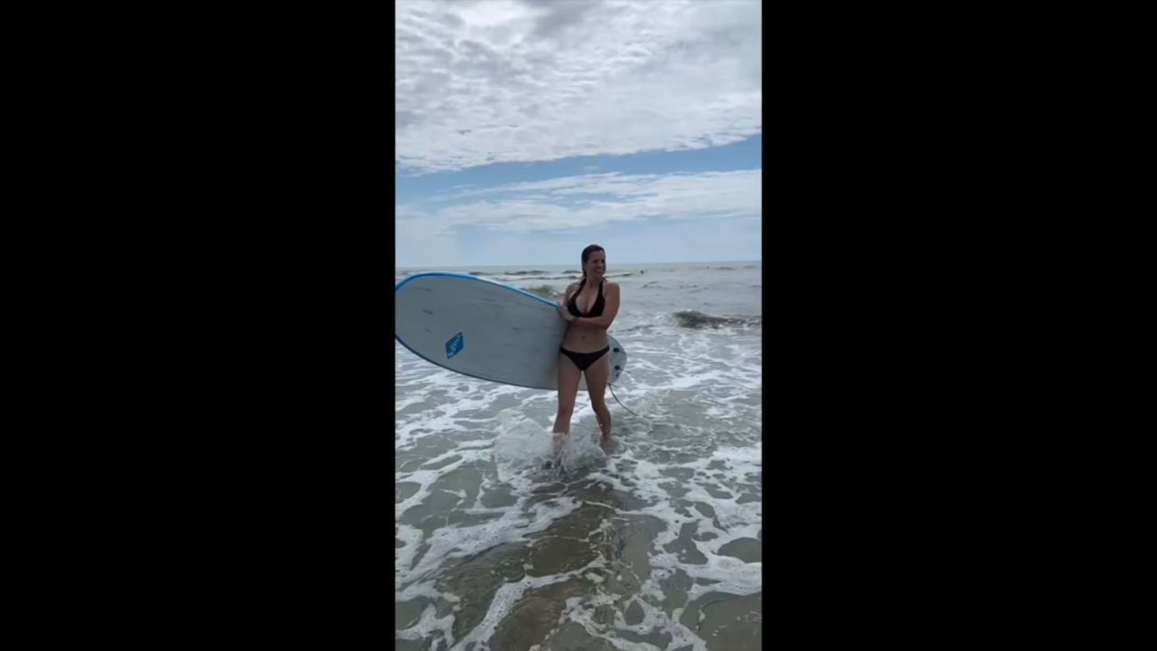 Beach swimsuit frame from the intro montage: Mace walking out of the surf carrying a surfboard