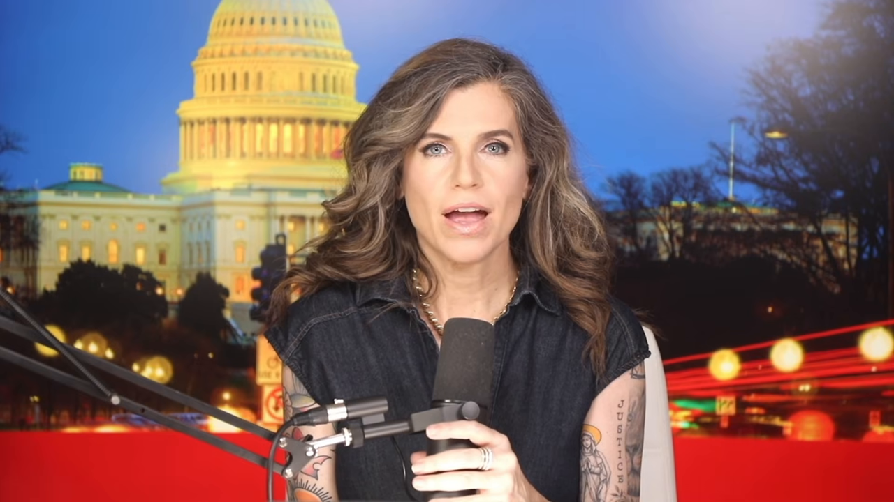 Studio-desk frame from a GET MACED episode: Nancy Mace at a boom microphone in a sleeveless dark denim top before a Capitol backdrop, her left forearm showing lettering and figure tattoos.
