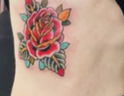 Close-up of a large American-traditional rose tattoo in full color, red bloom with green and teal leaves and orange accents, on Nancy Mace's left ribcage.